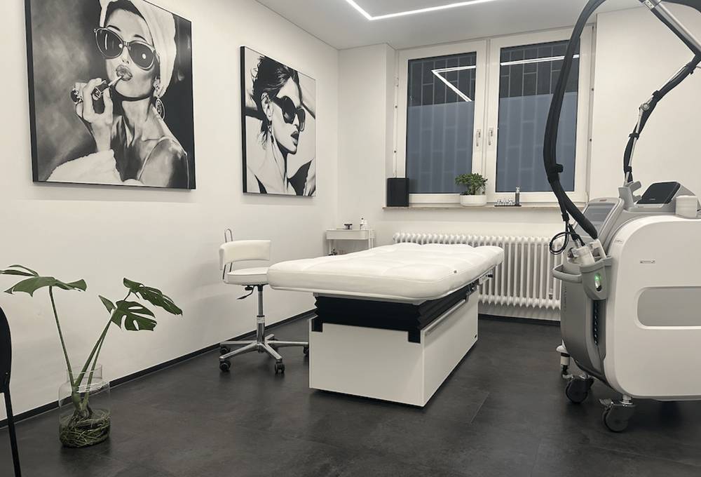 Behandlungsraum mit Liege, Hocker und Laser vor zwei Porträtbildern